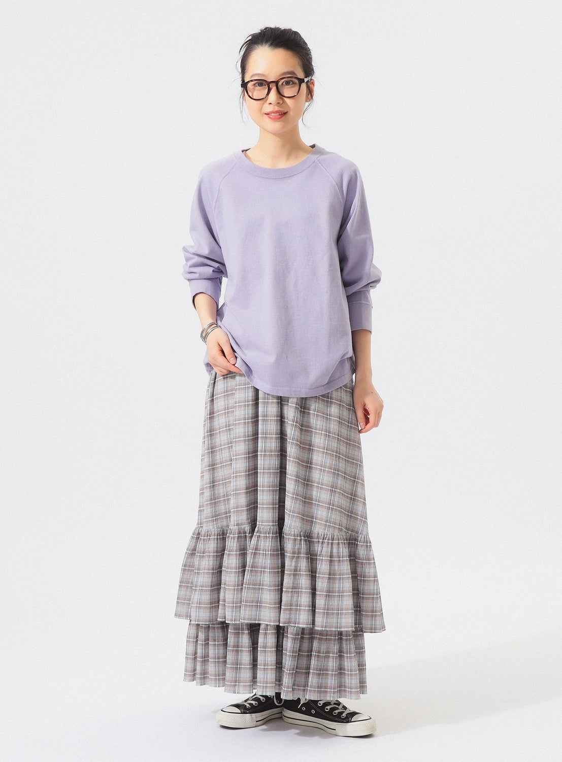 A woman wearing glasses, a light purple sweatshirt, the BEAMS BOY Check Tiered Skirt crafted from lightweight fabric in the brand’s signature check pattern, and black sneakers stands against a plain white background, smiling slightly.