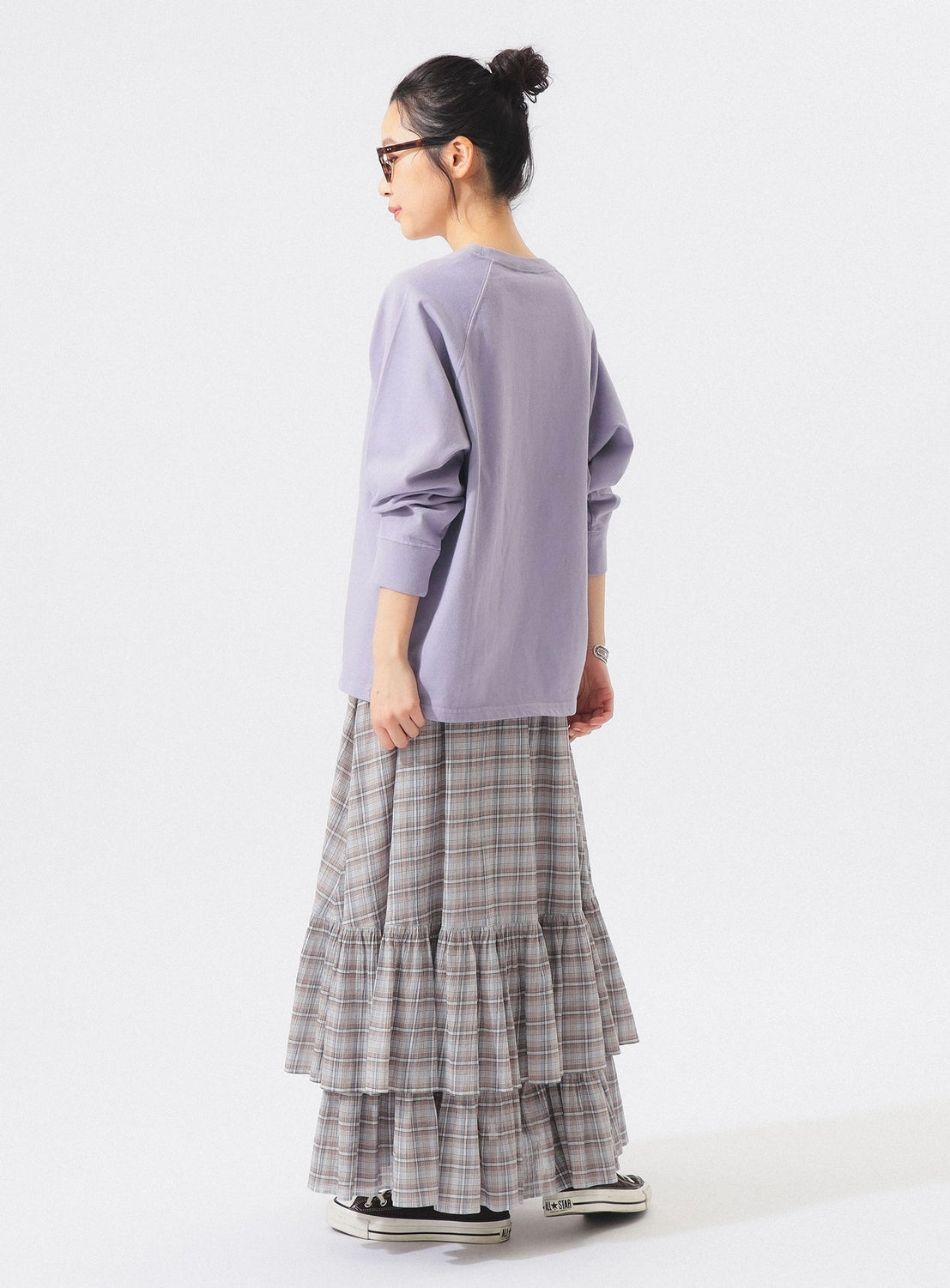 A woman with her hair in a bun, dressed in sunglasses, a loose lavender top, and sneakers stands facing away from the camera against a plain white background. She is wearing the BEAMS BOY Check Tiered Skirt, crafted from lightweight fabric featuring the brand’s signature check pattern.