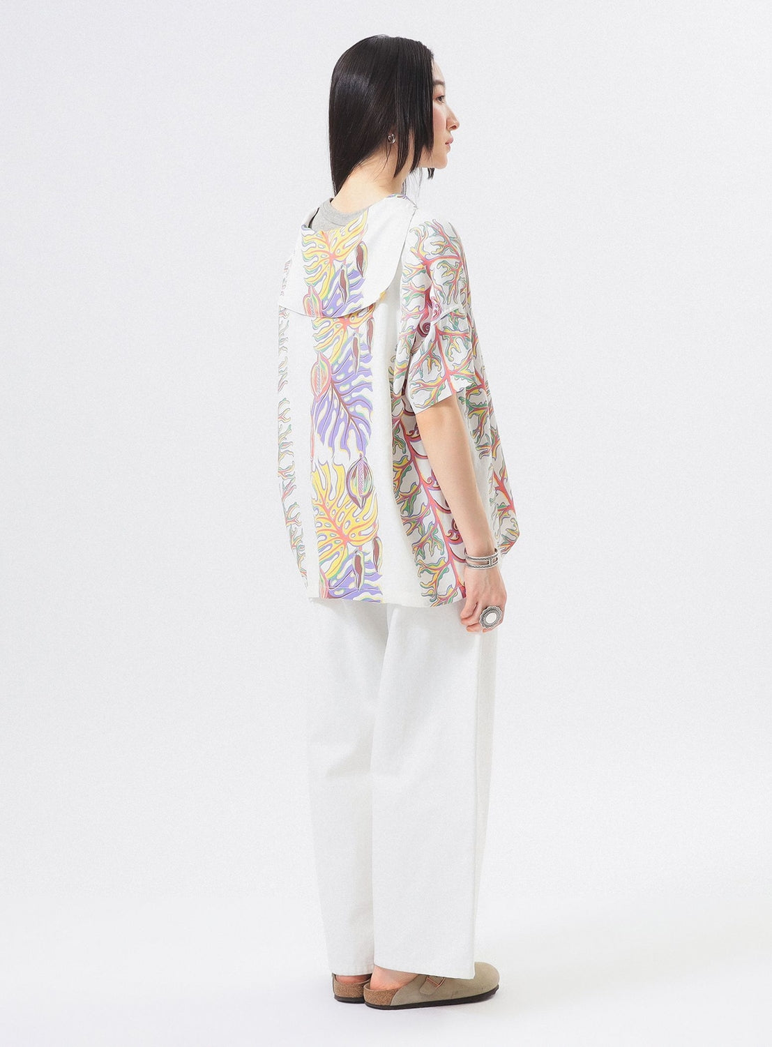 A woman with straight black hair wears the [Exclusive] SUN SURF x BEAMS BOY Sailor Shirt by SUN SURF, featuring colorful Hawaiian prints on the back and sleeves, paired with wide-leg pants and sandals, set against a plain white background.
