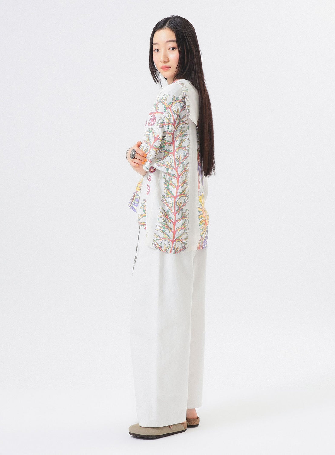 A woman with long dark hair stands sideways, looking at the camera. She wears the [Exclusive] SUN SURF x BEAMS BOY Sailor Shirt by SUN SURF, featuring colorful Hawaiian prints, with loose white pants and beige shoes against a plain white background.