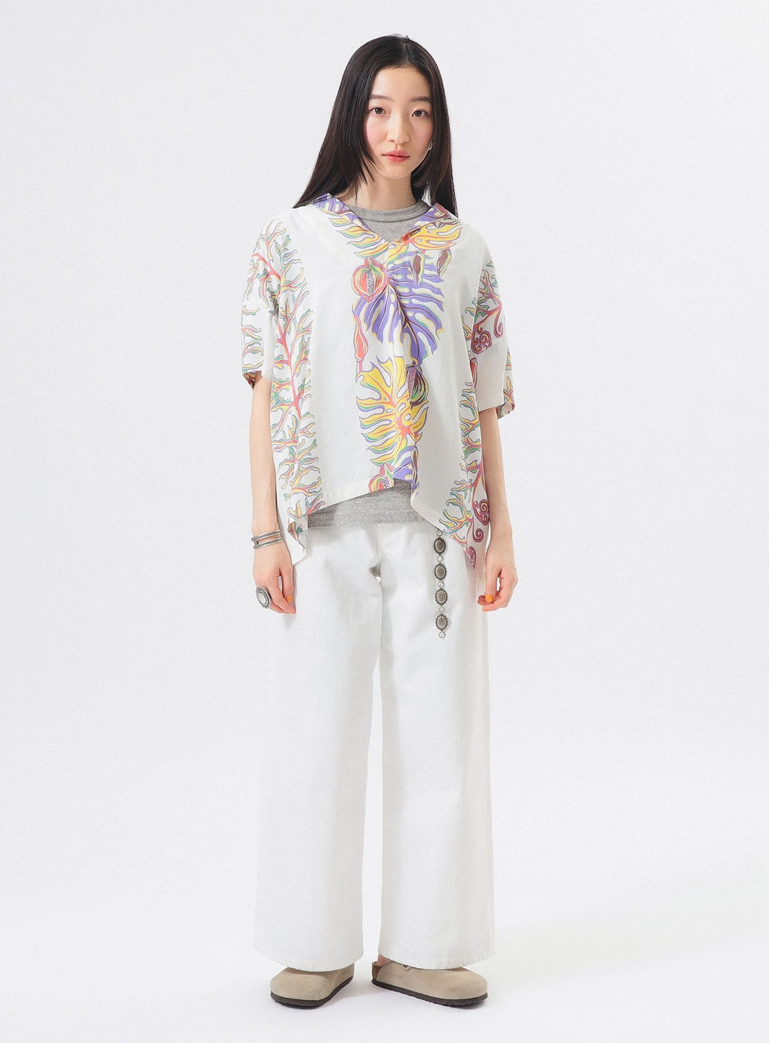A woman stands against a white background wearing the [Exclusive] SUN SURF x BEAMS BOY Sailor Shirt, featuring colorful Hawaiian prints, paired with wide-leg pants, beige shoes, and a scarf draped over her shoulders.