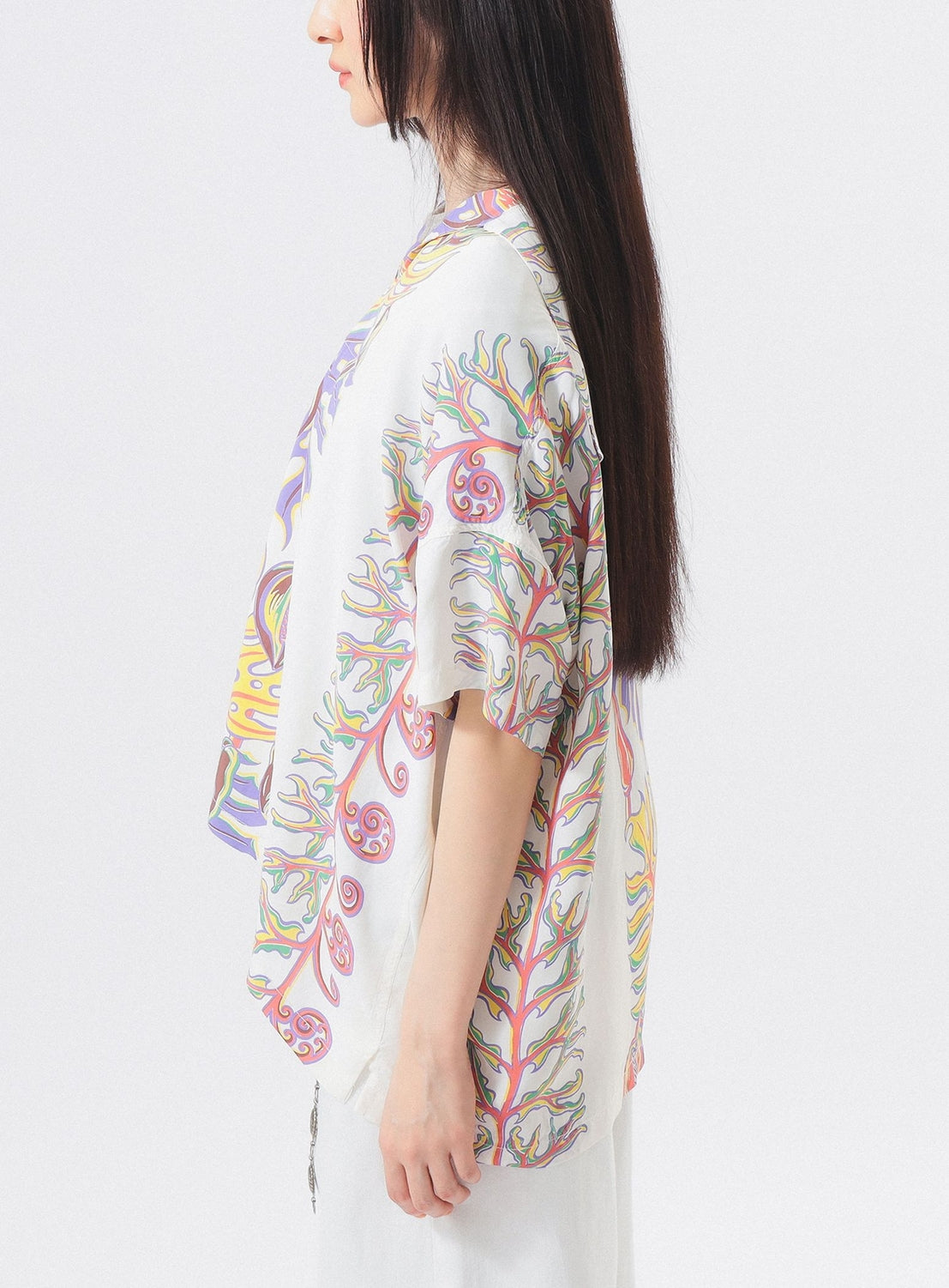 A woman with long straight hair stands sideways against a plain white background, wearing the [Exclusive] SUN SURF x BEAMS BOY Sailor Shirt by SUN SURF— a loose, short-sleeve white top with colorful Hawaiian-inspired wavy patterns and a white skirt.