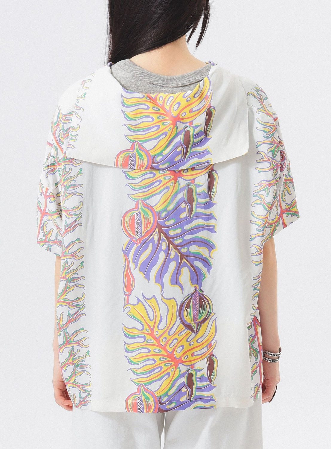 A person with long dark hair wears the [Exclusive] SUN SURF x BEAMS BOY Sailor Shirt, featuring colorful abstract Hawaiian prints on the back and sleeves. Shown from behind; a standout piece by SUN SURF.