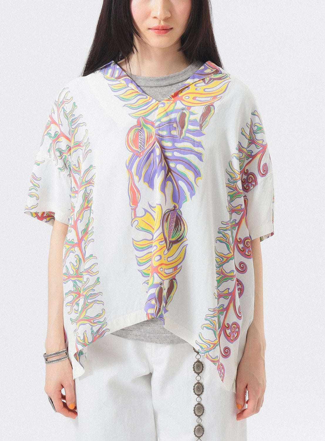 A woman wears the [Exclusive] SUN SURF x BEAMS BOY Sailor Shirt by SUN SURF, featuring colorful Hawaiian-inspired abstract leaf patterns, paired with white pants with decorative buttons. Her face is partially out of frame and her dark hair is down.
