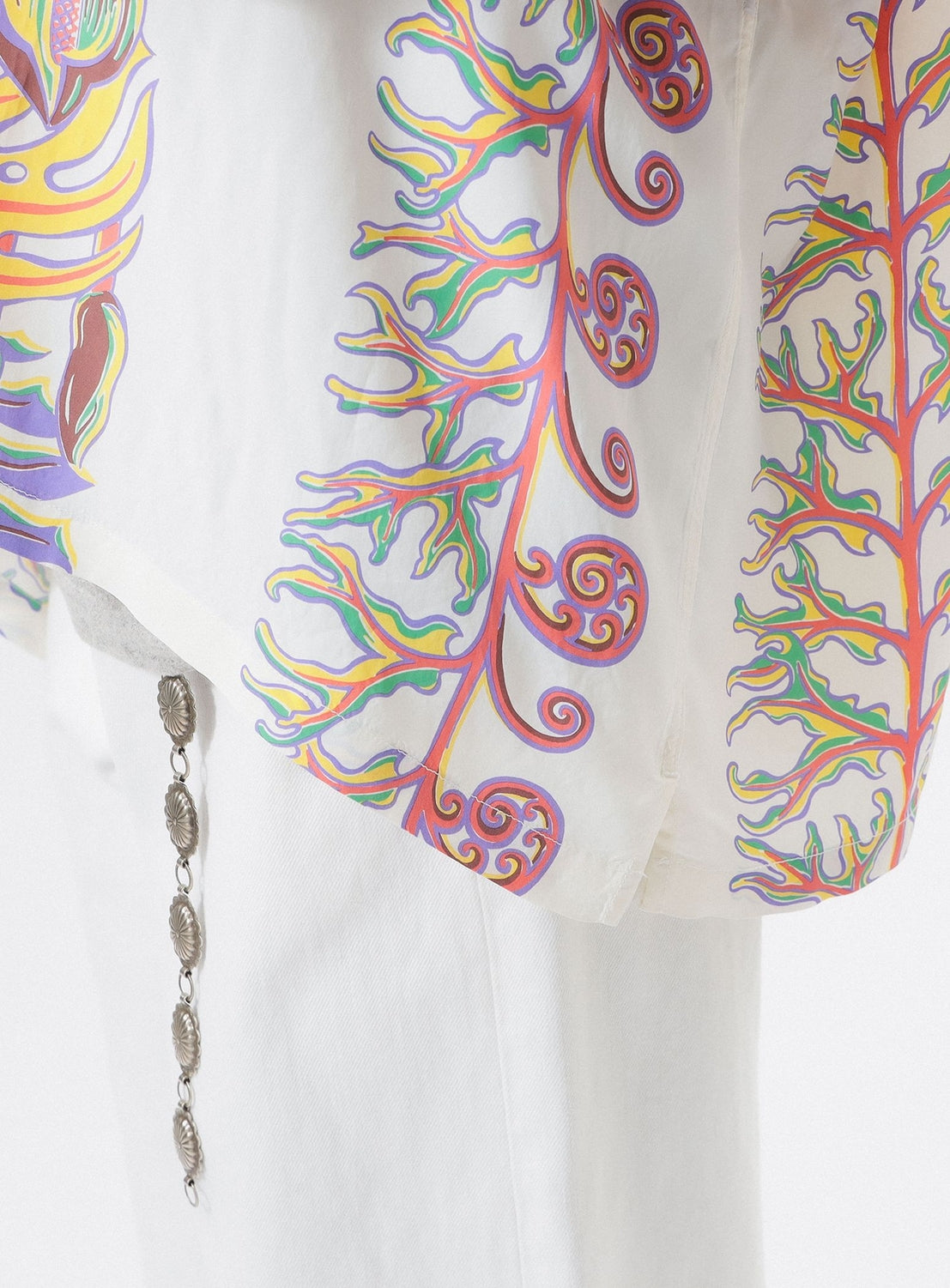 A close-up of the [Exclusive] SUN SURF x BEAMS BOY Sailor Shirt by SUN SURF, featuring sheer white fabric with colorful Hawaiian-inspired botanical prints, draped over white pants and styled with a silver oval-charm chain.