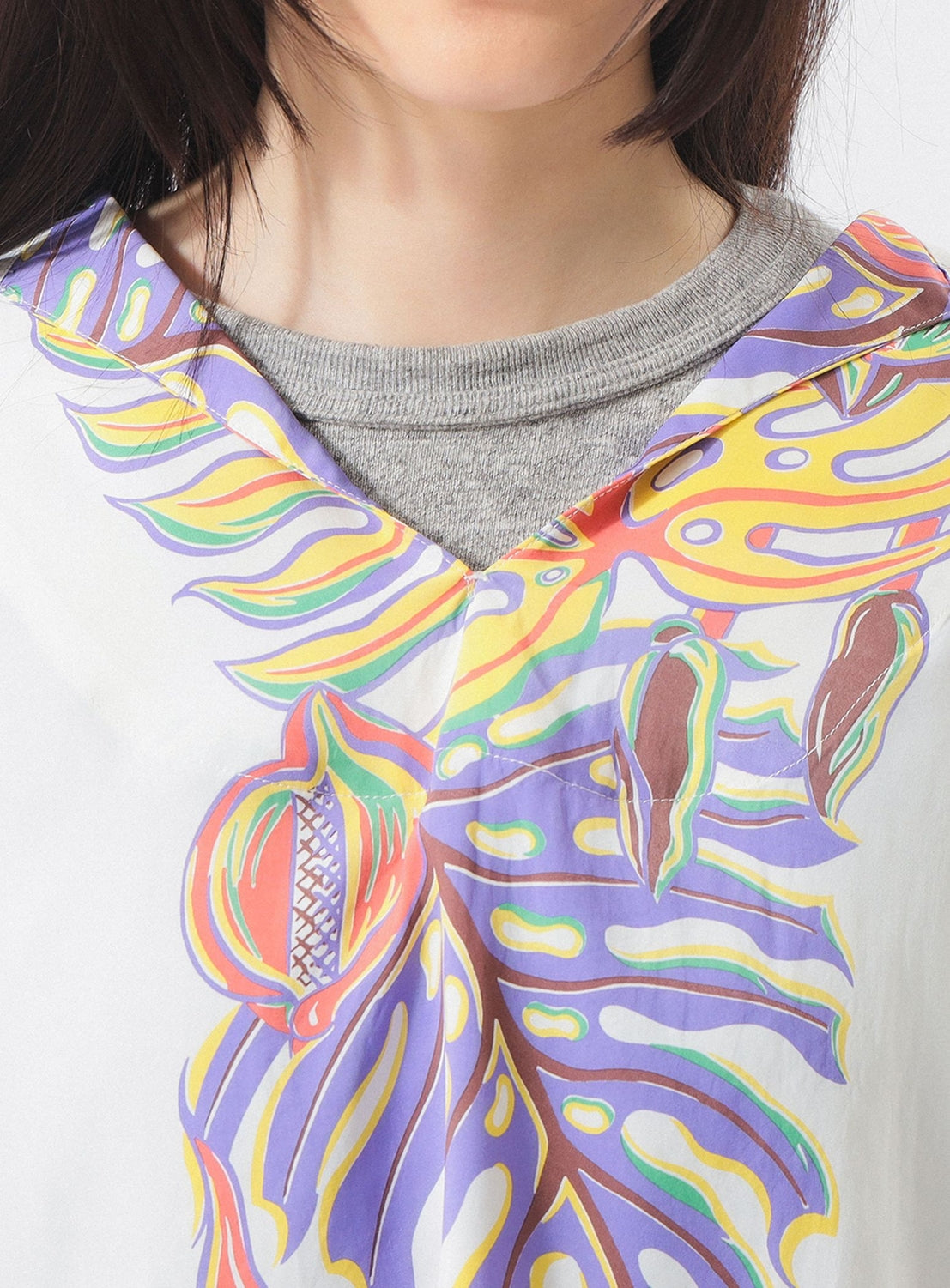 A close-up shows a person wearing the [Exclusive] SUN SURF x BEAMS BOY Sailor Shirt—featuring colorful Hawaiian prints by SUN SURF—over a gray crew-neck top. Only the lower half of their face and neck are visible.