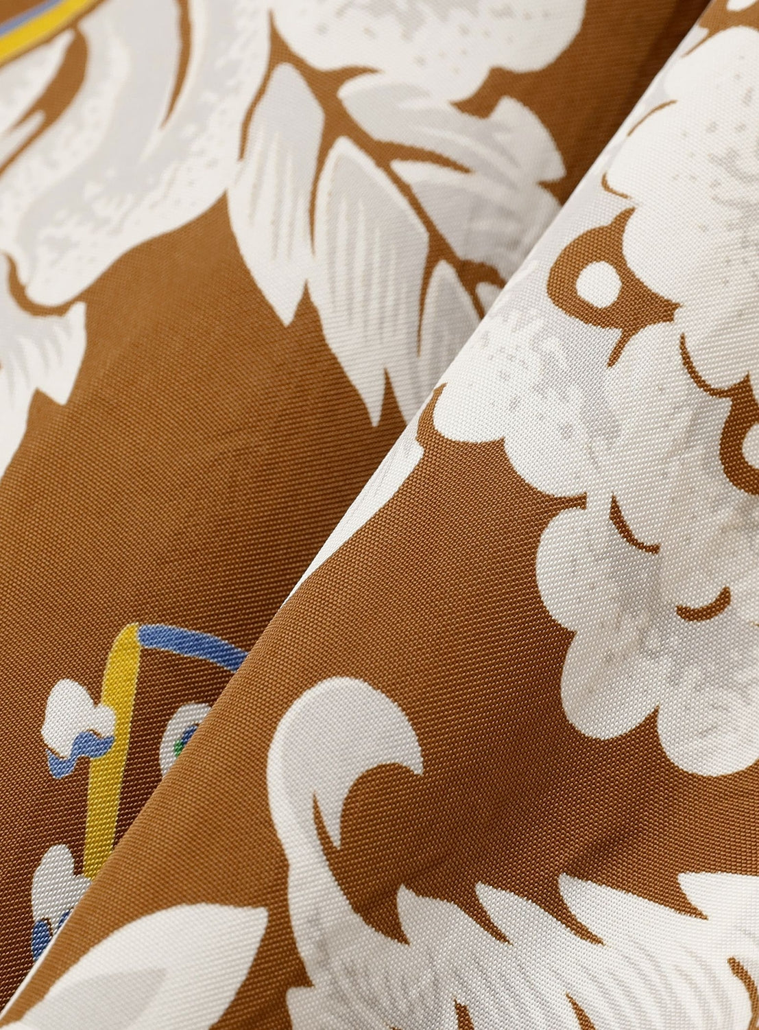 Close-up of SUN SURF fabric with a brown background, white floral patterns, and yellow-blue accents. The soft creases evoke resort vibes—ideal for the [Exclusive] SUN SURF Sleeveless Cami Dress, inspired by vintage aloha style.