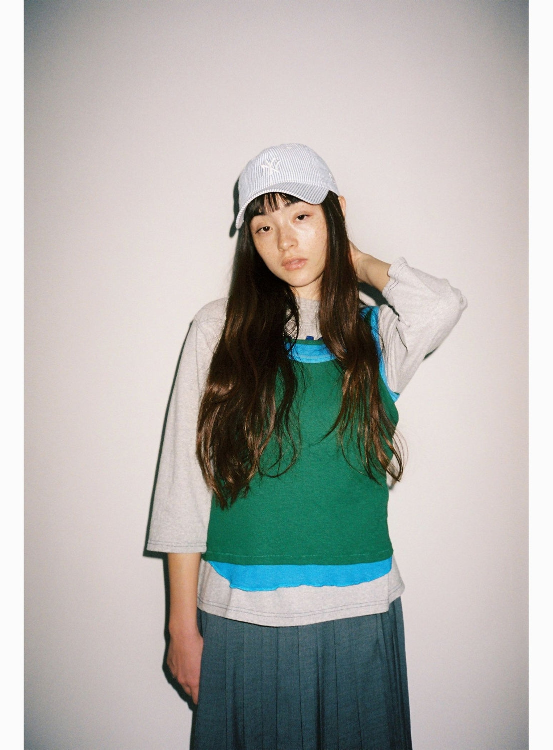 A young woman with long brown hair wears a white cap, a pleated skirt, and the BEAMS BOY Double-Layered Sheer Ribbed Tank Top in green and gray, standing against a plain white background with one hand behind her head.