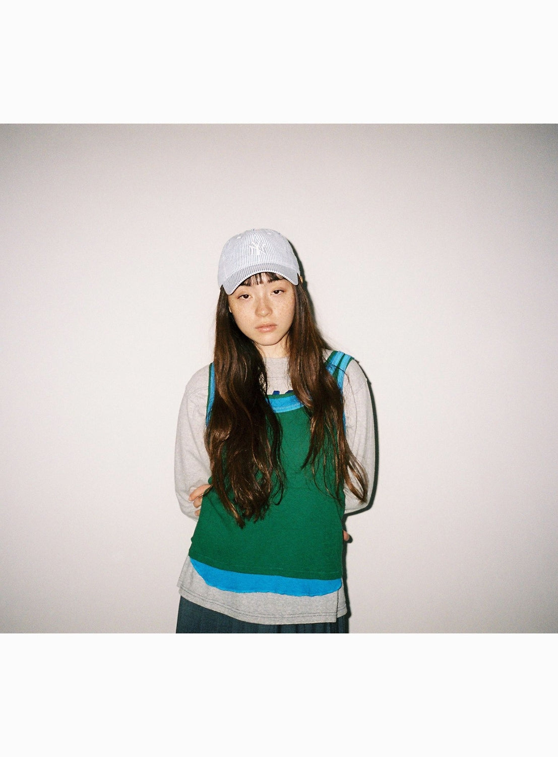 A person with long brown hair, wearing a white cap and the BEAMS BOY Double-Layered Sheer Ribbed Tank Top, stands against a plain white wall, looking at the camera with their arms behind their back.