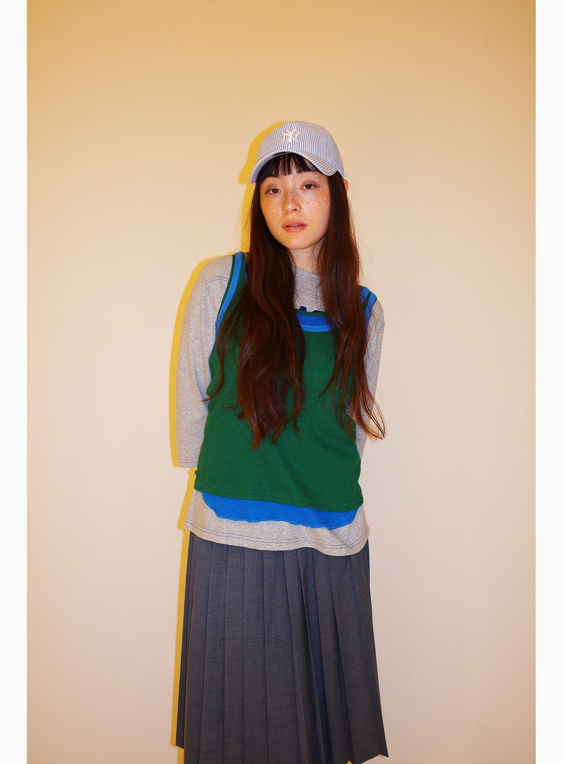 A young woman with long brown hair stands against a plain beige background, wearing a gray cap, the BEAMS BOY Double-Layered Sheer Ribbed Tank Top in green and blue over a gray shirt, and a gray pleated skirt.