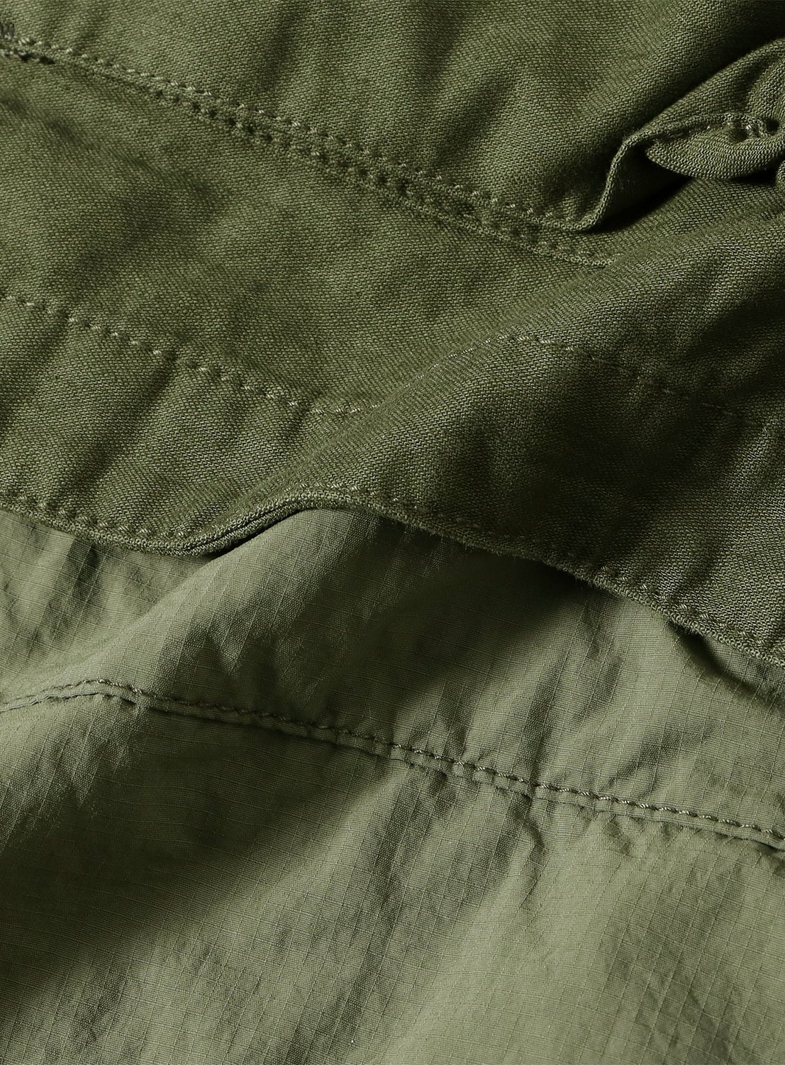 Close-up of olive green fabric with visible seams and stitching highlights the texture and natural folds of the material. The fabric appears thick and sturdy, characteristic of a BEAMS Wide Fit M-65 Layered Jacket, and showcases features such as a double-zip front and an adjustable silhouette inspired by classic military outerwear.