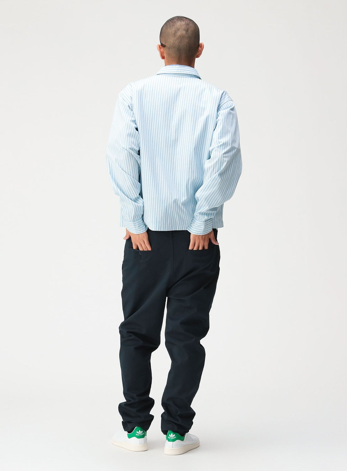 A person seen from behind, wearing a light blue and white striped Striped Zip-Up Shirt and dark trousers, with their hands in their pockets.