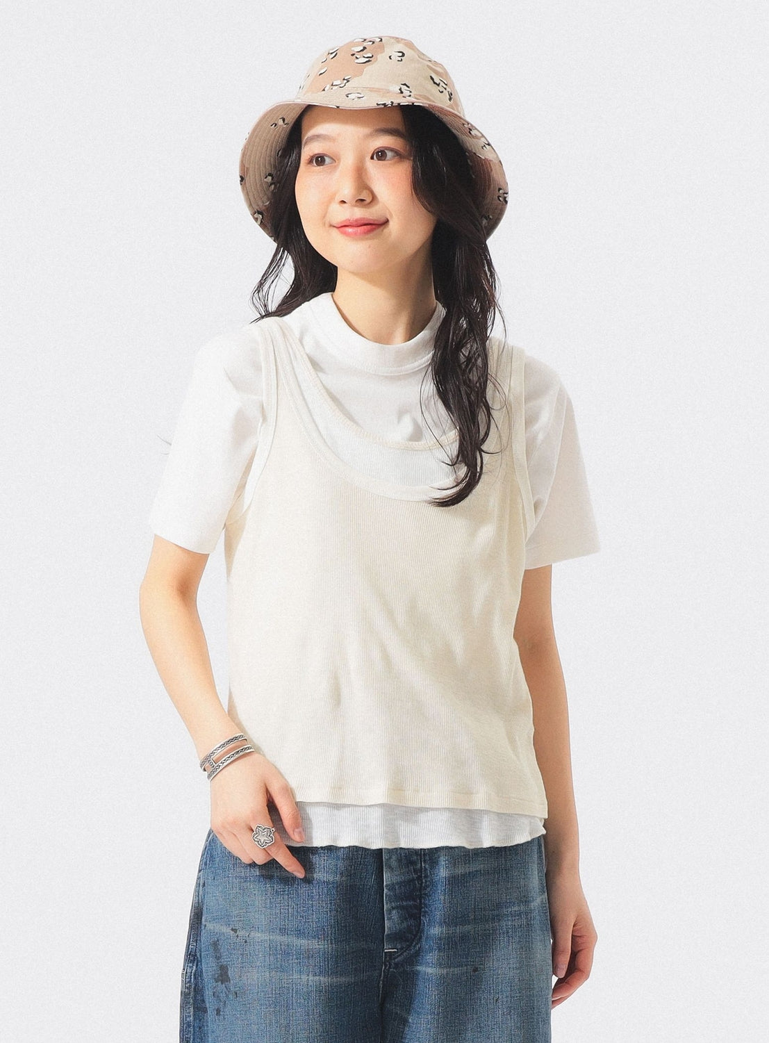 A woman stands against a plain white background wearing the BEAMS BOY Layered Rib Tank Top—a cream, breathable cotton piece styled over a white top—paired with blue jeans, bracelets, and a beige bucket hat featuring a cartoon pattern.