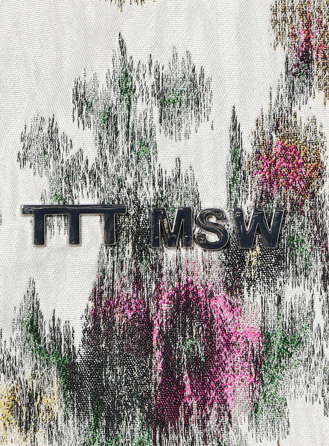 Black metal letters spelling TTT MSW rest on Italian-made fabric echoing the green, pink, and gold abstract floral patterns of the Flower Jacquard Anorak Hoodie by TTT MSW.