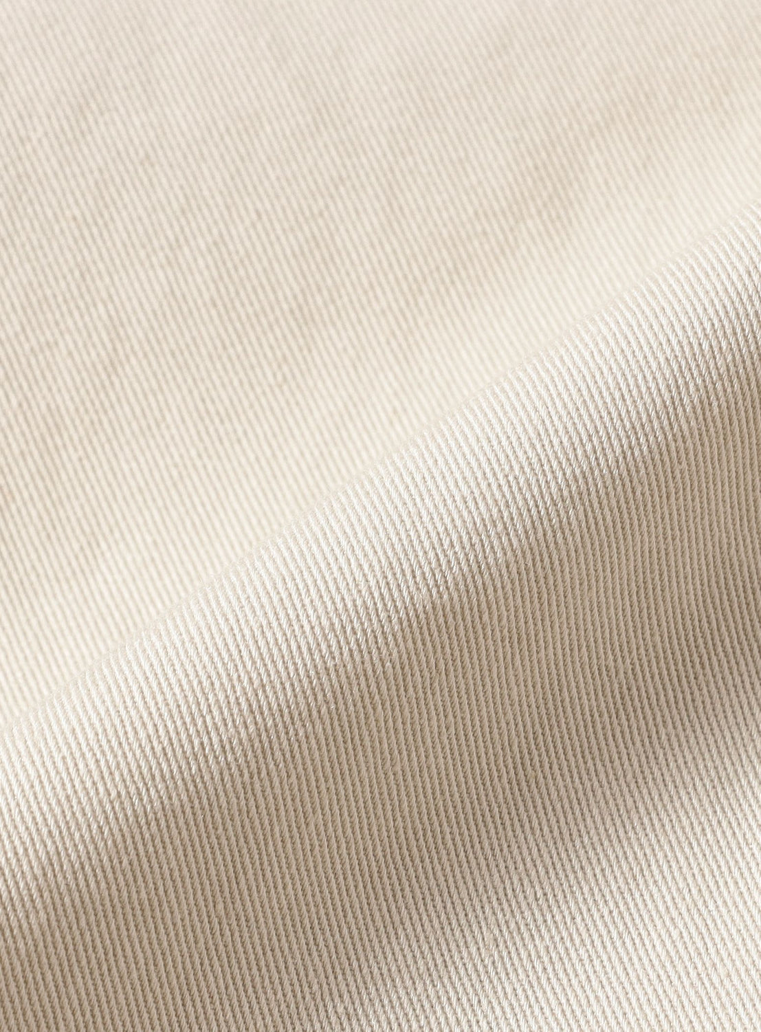 Close-up of cream-colored, ribbed fabric with diagonal textured lines and soft folds, highlighting the distinctive weave pattern and gentle shadows—an ideal choice for BEAMS’ Basic Twill Easy Pants, which offer relaxed comfort and effortless style.