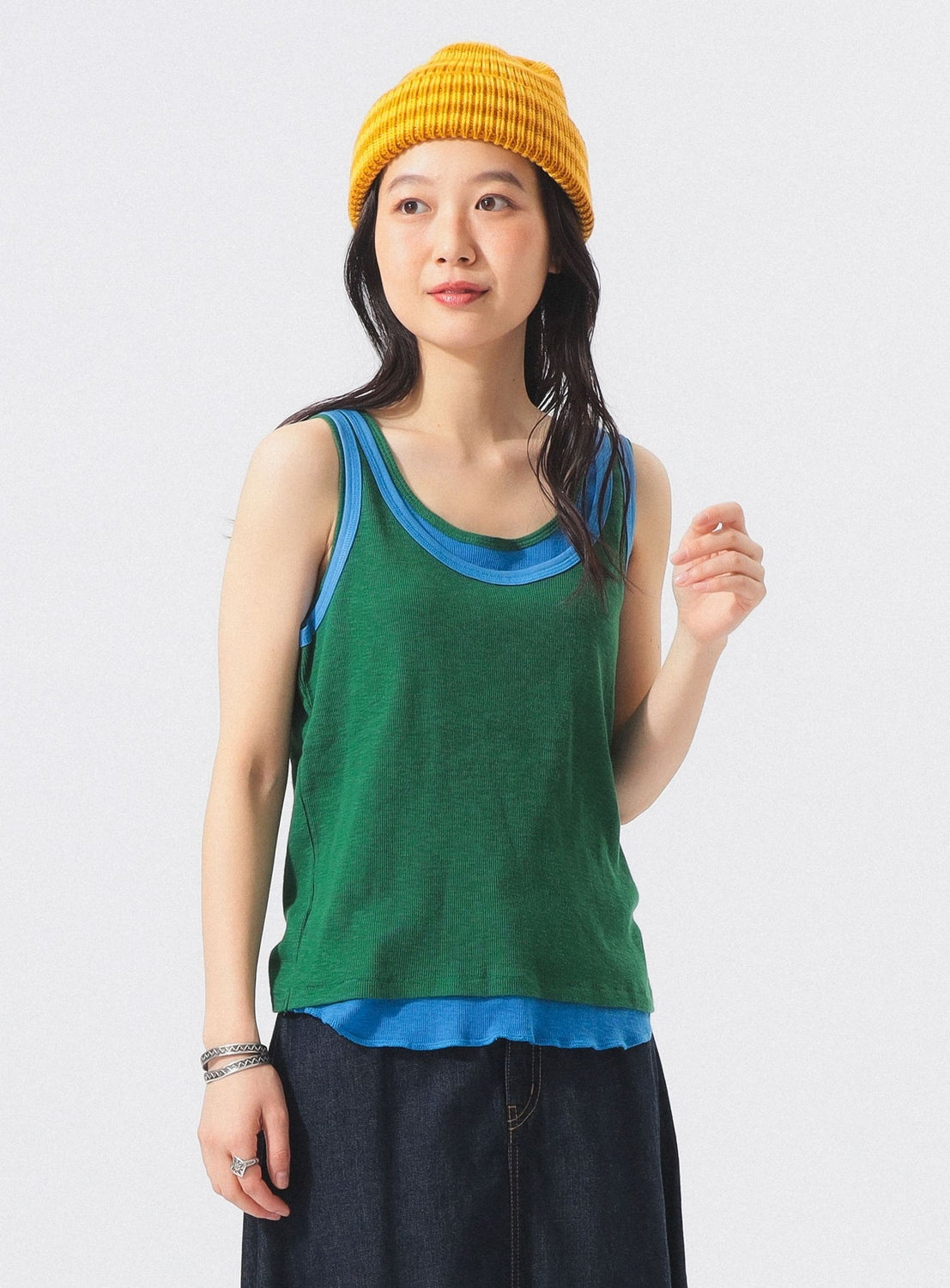 A person stands against a plain white background, looking slightly to the side with a soft smile, wearing a yellow ribbed knit beanie, a green sleeveless top layered over the BEAMS BOY Double-Layered Sheer Ribbed Tank Top in blue, paired with a dark skirt.