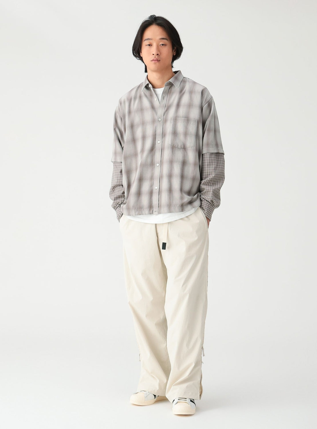 A man wears a grey, faux-layered BEAMS Layered Check Shirt with a plaid pattern, a white undershirt, and wide-fit, off-white trousers.