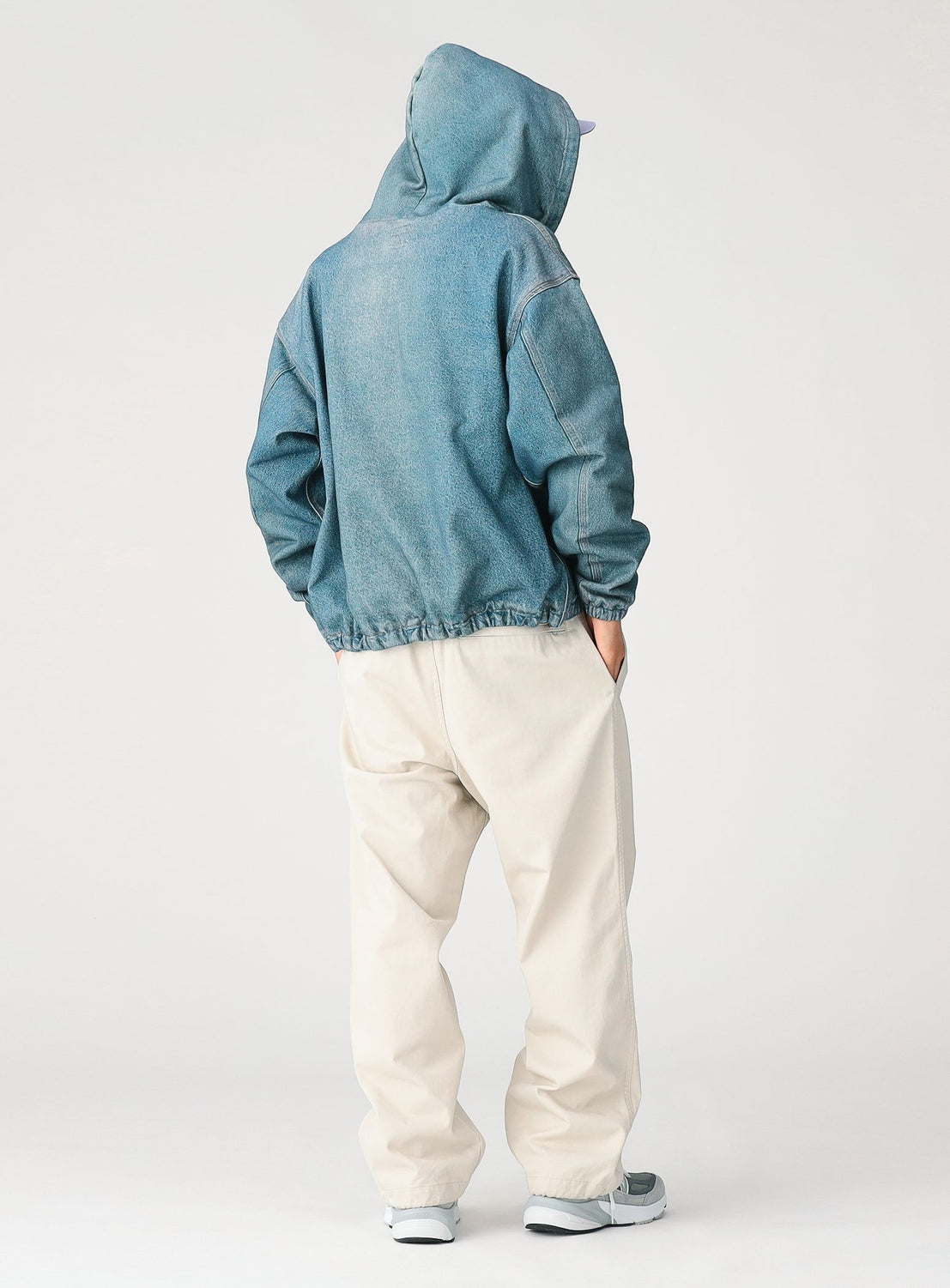 A person stands facing away, wearing a blue denim hoodie, BEAMS Basic Twill Easy Pants in light beige with a relaxed fit, and white sneakers. Their hands are in their pockets and the hood is up, covering their head. The background is plain white.