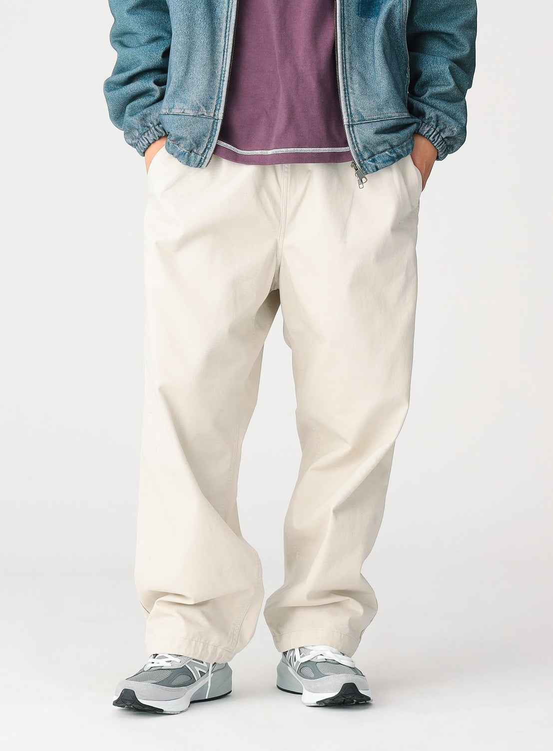 A person stands with hands in pockets, wearing BEAMS Basic Twill Easy Pants in off-white—a relaxed fit cotton twill style featuring an adjustable waistband—paired with gray sneakers, a purple shirt, and a blue denim jacket. The image is cropped to show only from the shoulders down.