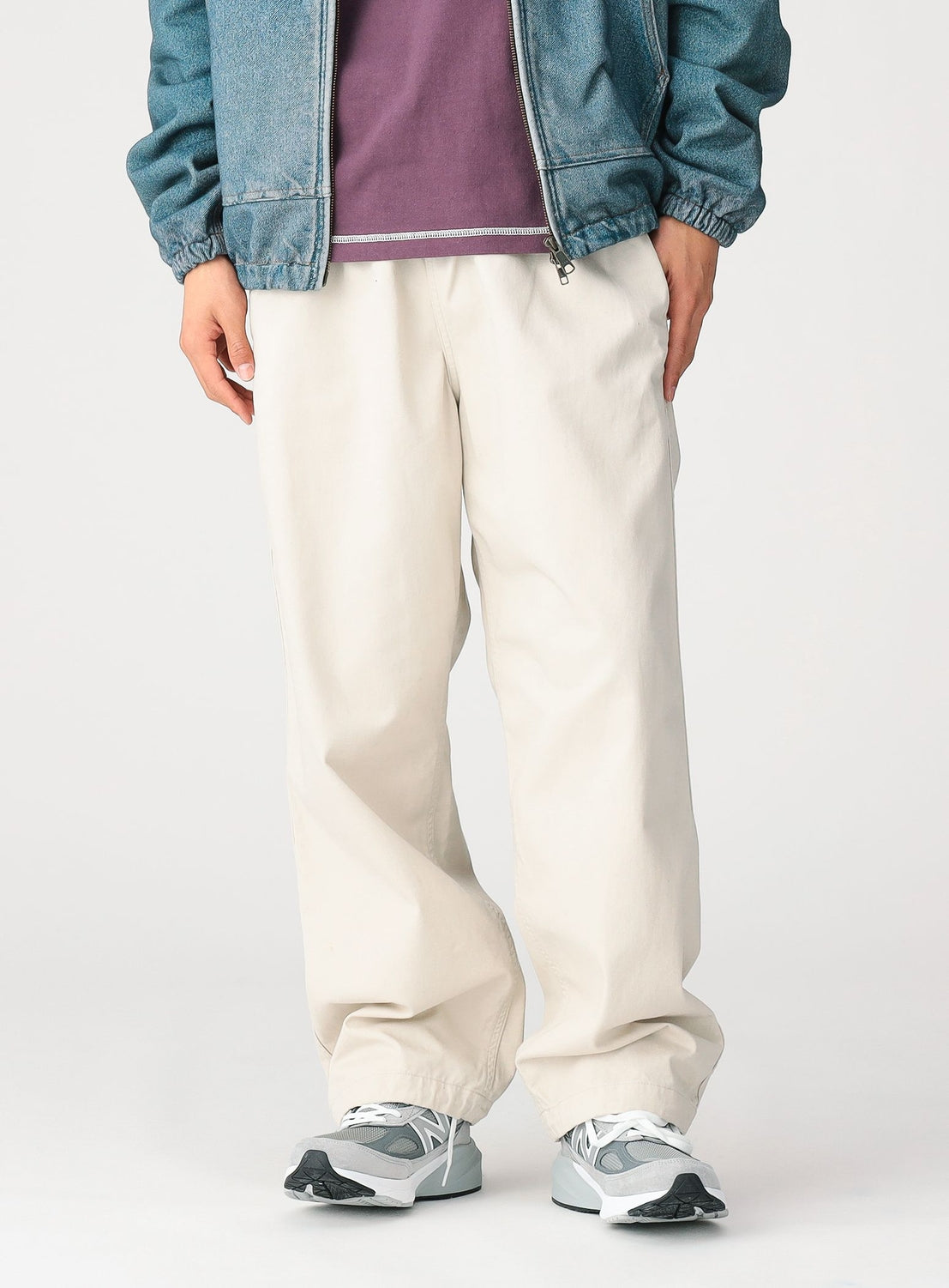 A person wearing BEAMS Basic Twill Easy Pants in a loose, relaxed cream-colored silhouette with an adjustable waistband, paired with gray and white sneakers, a purple shirt, and a blue zip-up jacket. The photo is cropped at the shoulders.