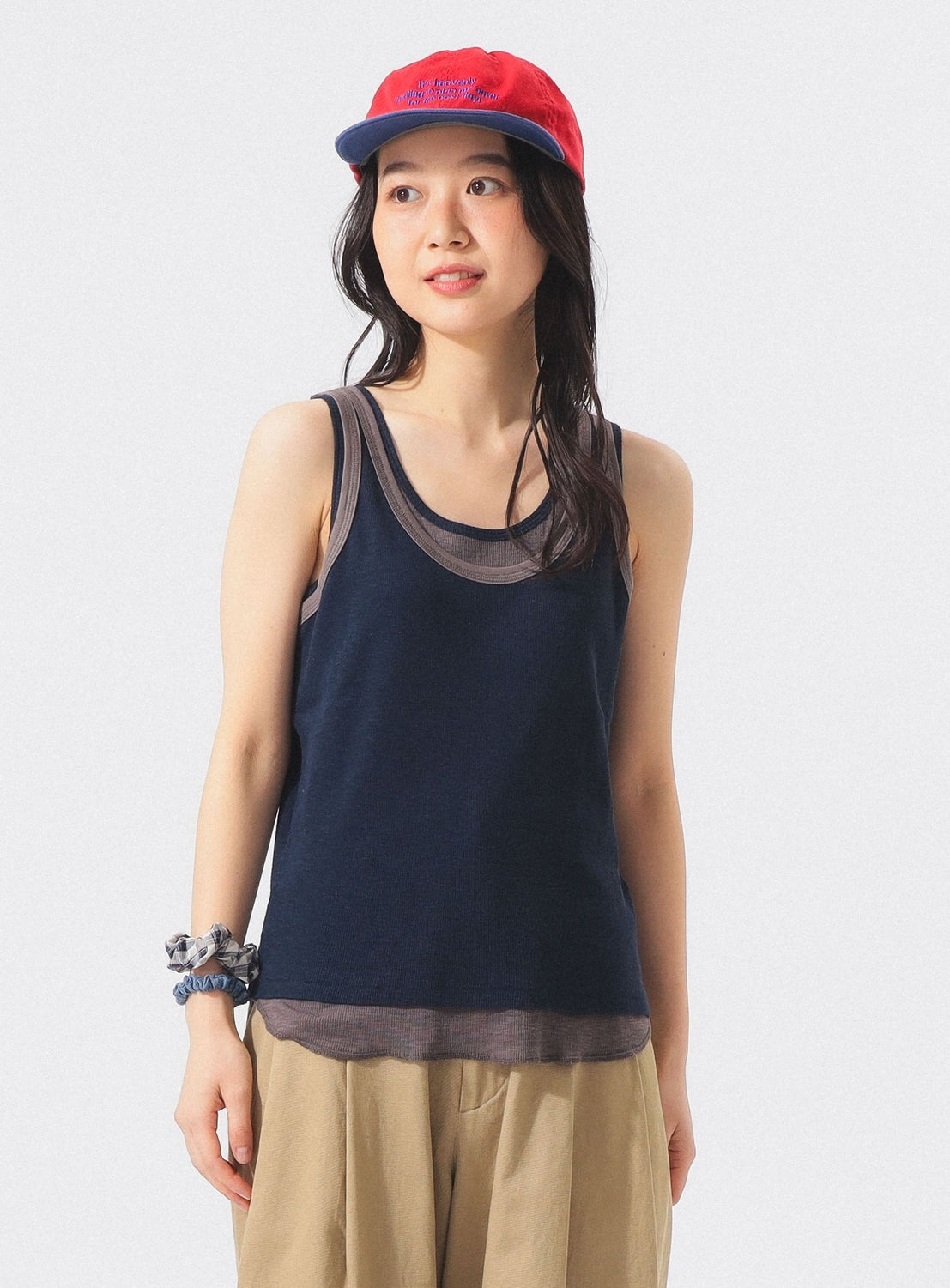 A woman with long dark hair and a scrunchie on her wrist stands against a plain light background, looking slightly to the side. She is wearing the BEAMS BOY Layered Rib Tank Top in navy, layered over a sheer brown top, paired with beige pants and a red cap.