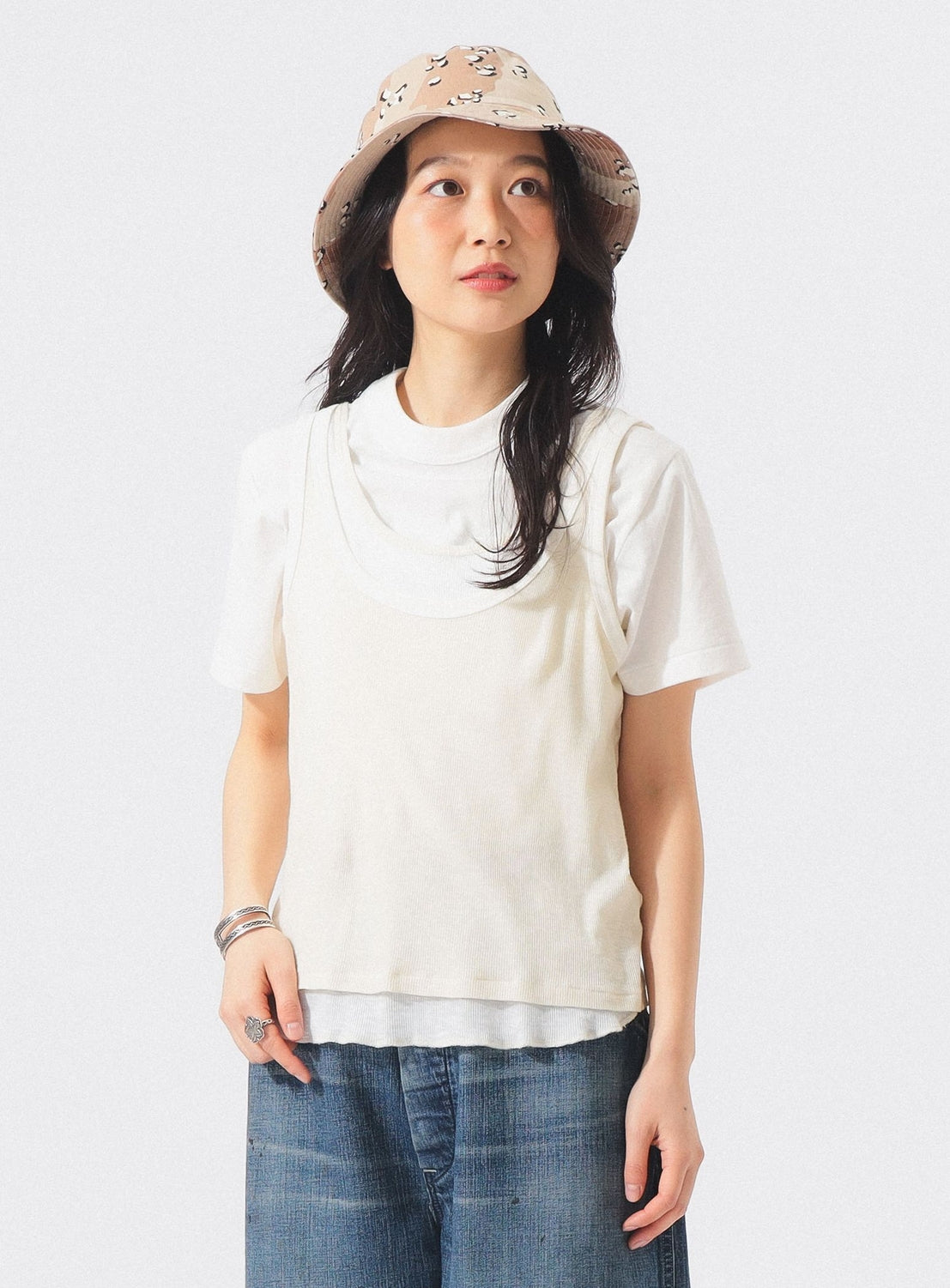 Wearing the BEAMS BOY Layered Rib Tank Top, a woman in a beige hat with black patterns and blue jeans stands against a plain light background, gazing to her left.