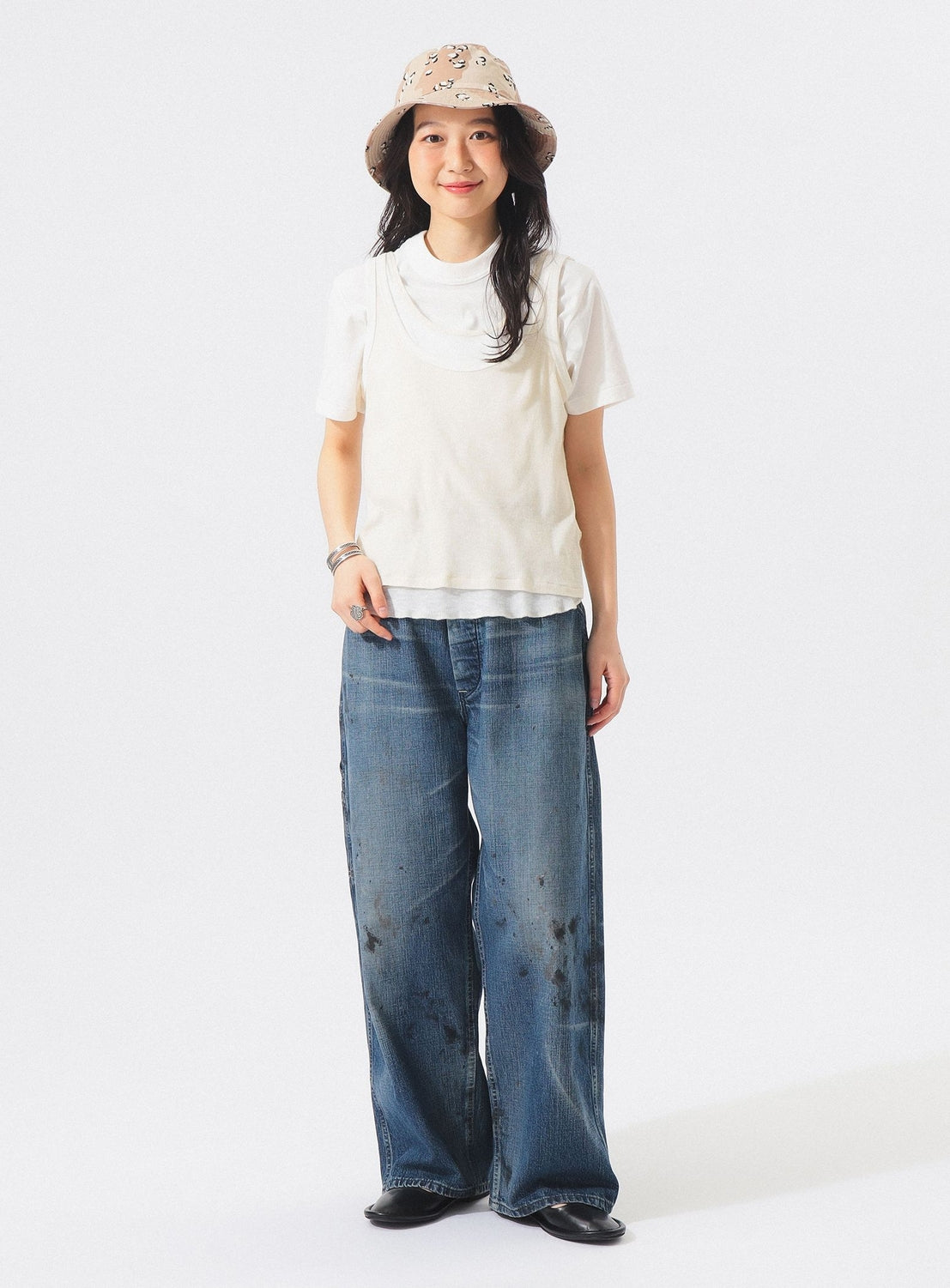 A person stands smiling against a plain white background, wearing the BEAMS BOY Layered Rib Tank Top, a beige hat, and wide-leg blue jeans with dark stains.