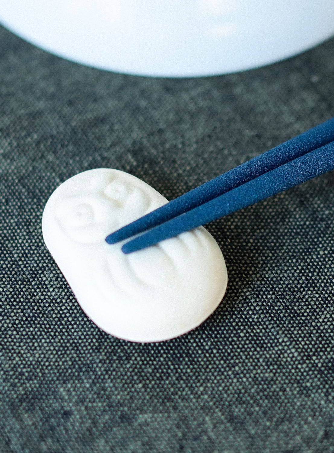 White daruma chopstick rest with blue chopsticks on a textured dark surface