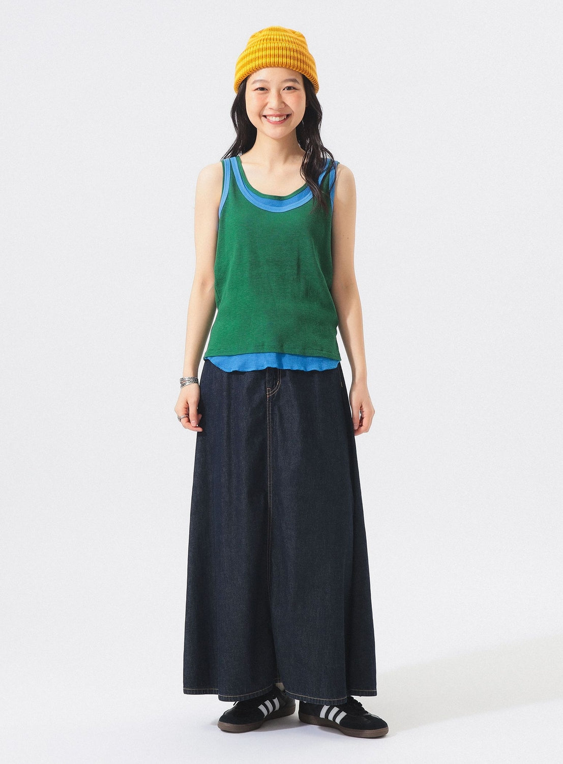 A woman wearing a yellow beanie, a BEAMS BOY Double-Layered Sheer Ribbed Tank Top over a blue shirt, a long denim skirt, and black shoes stands smiling against a plain white background.