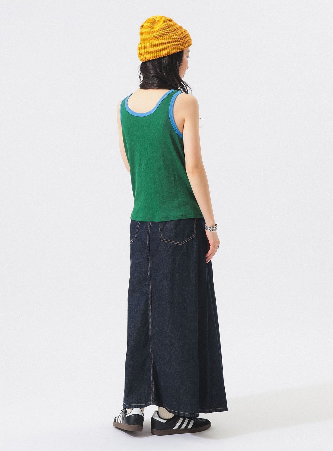 A person faces away, wearing a yellow knit beanie, the BEAMS BOY Double-Layered Sheer Ribbed Tank Top—a 100% cotton tank with blue trim—a long dark denim skirt, black and white sneakers, and bracelets, standing against a plain white background.