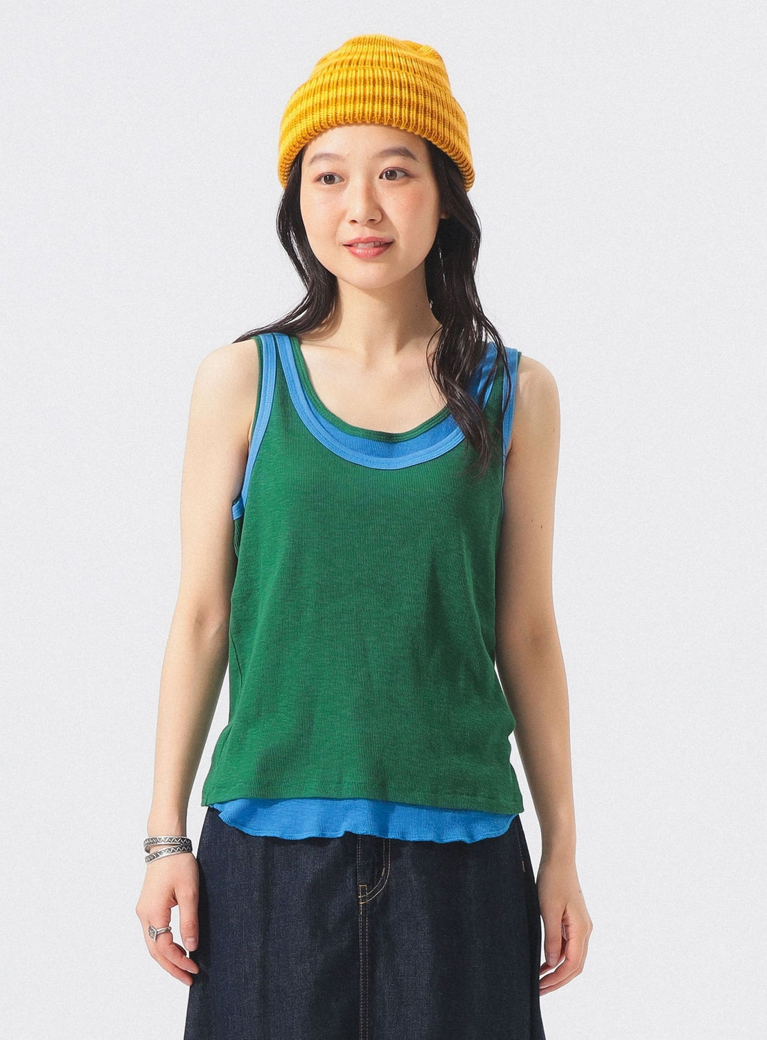 A person stands against a plain light background, smiling slightly, dressed in a yellow knit hat and dark pants, and wearing the BEAMS BOY Double-Layered Sheer Ribbed Tank Top—a green tank top layered over a blue ribbed knit tank.