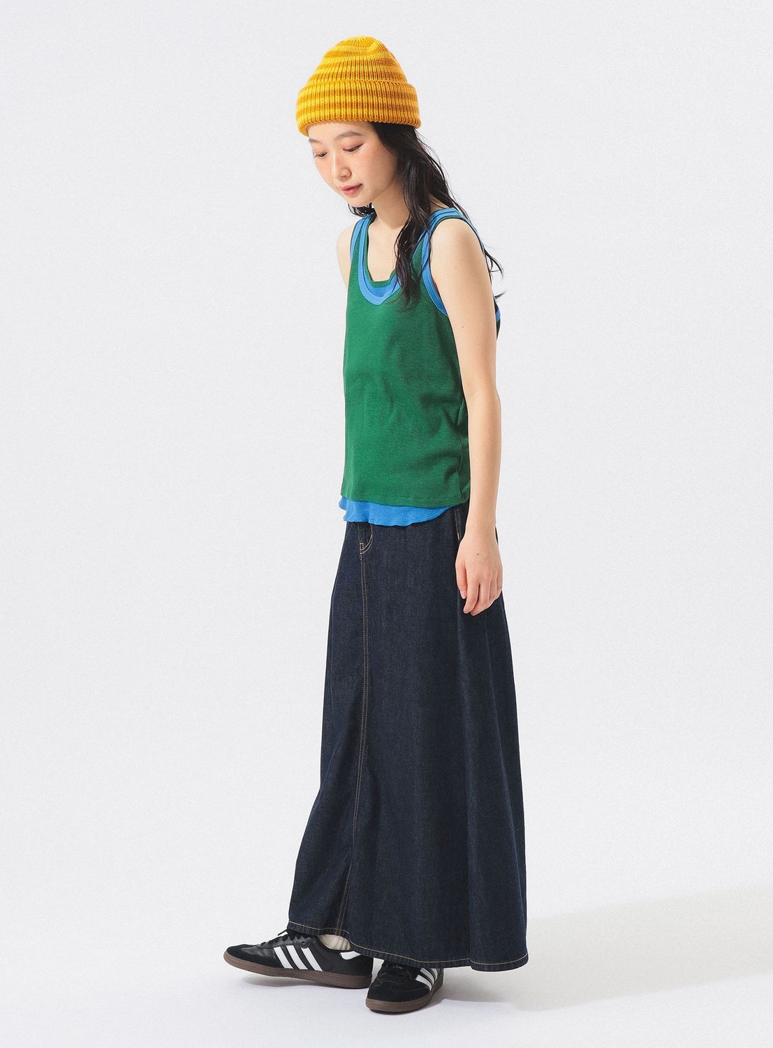 A person wearing a yellow beanie, a BEAMS BOY Double-Layered Sheer Ribbed Tank Top in green over a blue shirt, a long dark skirt, and black Adidas sneakers stands against a plain white background.