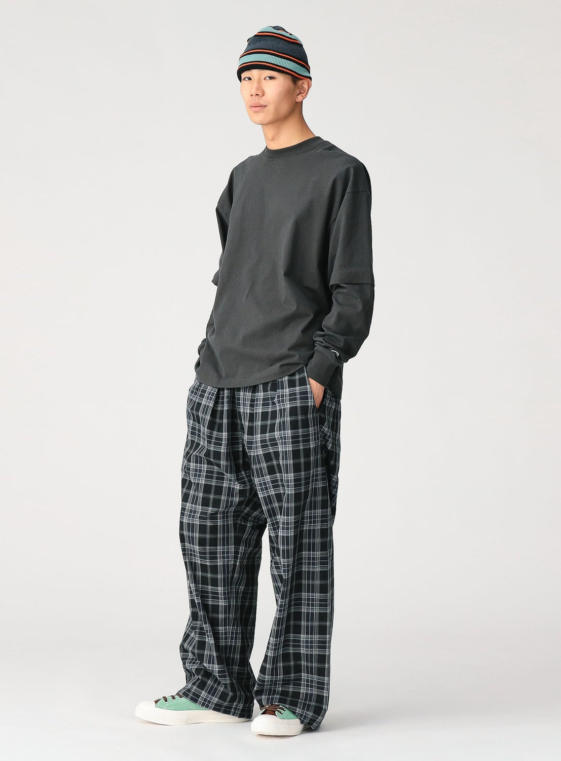 A person stands against a plain background wearing a striped beanie, CHAMPION’s [Exclusive] Mock Layered Long-Sleeve T-Shirt, wide plaid pants, and white sneakers with green accents. Their hands are in their pockets and they look relaxed.