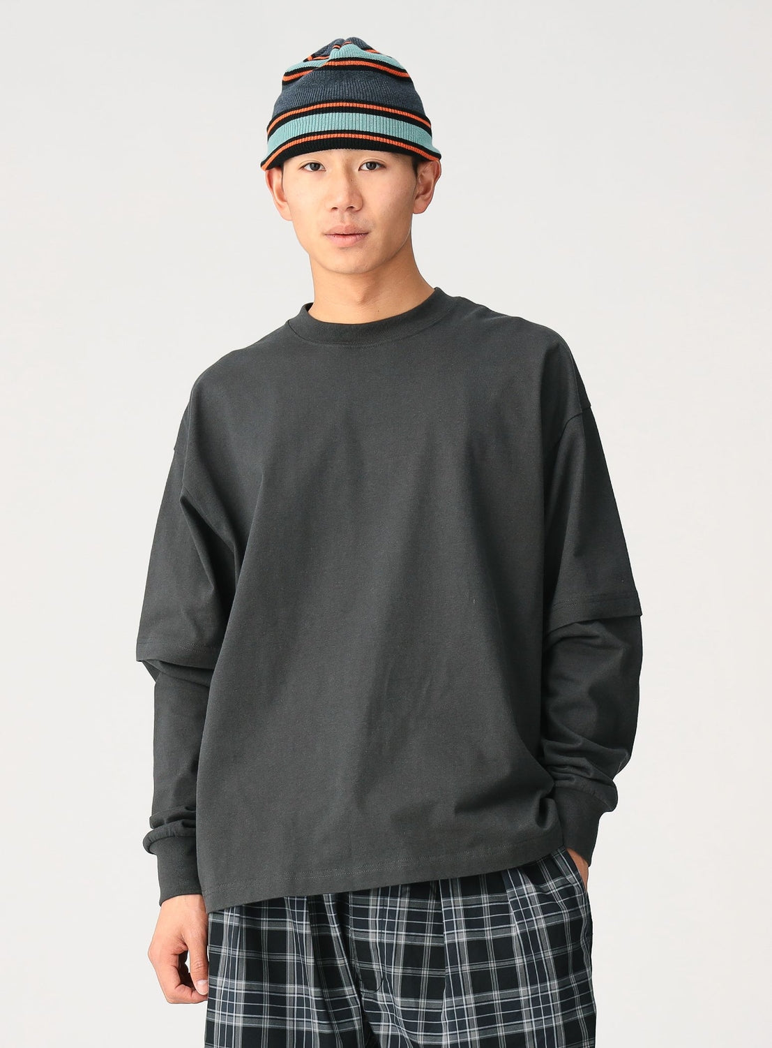 A young person stands against a plain background wearing a black [Exclusive] Mock Layered Long-Sleeve T-Shirt from CHAMPION, plaid pants, and a striped beanie. They face the camera with a neutral expression.