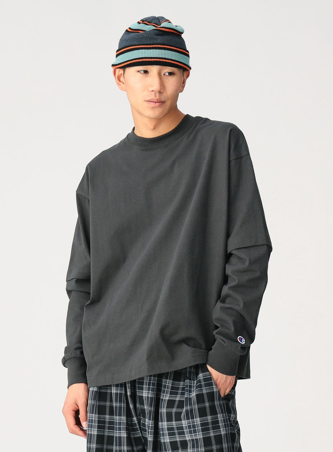 Wearing a CHAMPION [Exclusive] Mock Layered Long-Sleeve T-Shirt, plaid pants, and a colorful knit beanie, a person stands against a plain light background with a neutral expression, highlighting the shirt’s comfortable fit.