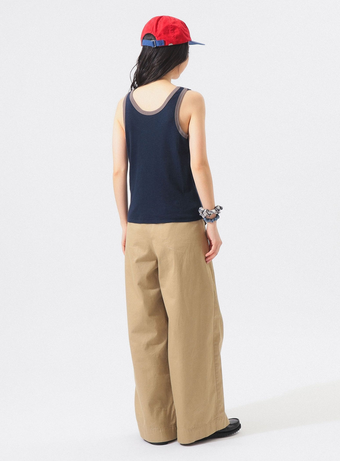 A person stands facing away against a plain white background, wearing a red cap, BEAMS BOY Layered Rib Tank Top in navy, tan wide-leg pants, black shoes, and a scrunchie on one wrist.