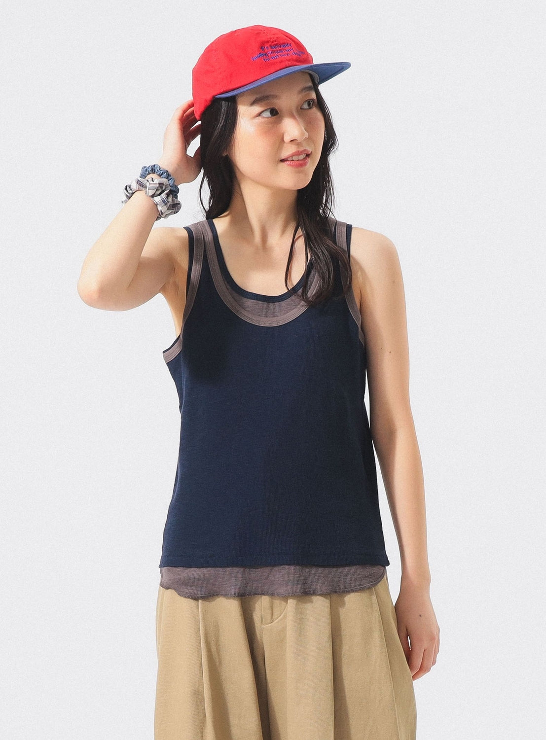A woman wearing the BEAMS BOY Layered Rib Tank Top in navy, a red cap, and beige pants stands against a plain background, touching her cap with one hand and looking to the side.
