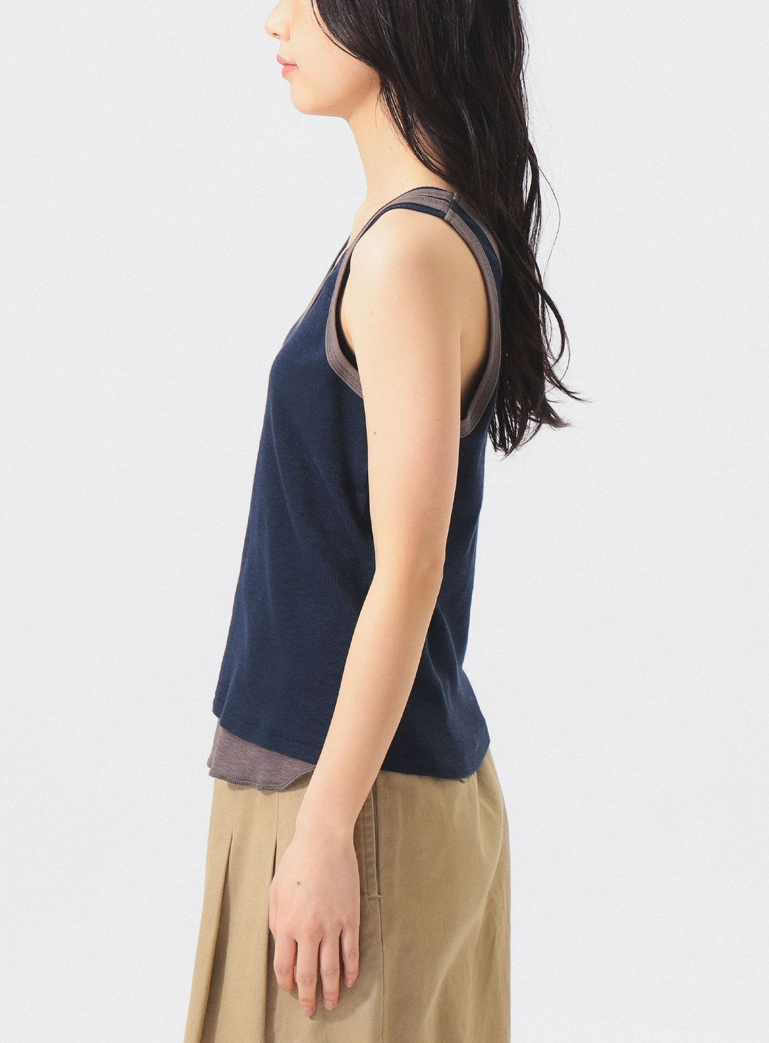 A woman with long dark hair stands in profile against a plain light background, wearing the BEAMS BOY Layered Rib Tank Top over a gray top and a beige skirt.