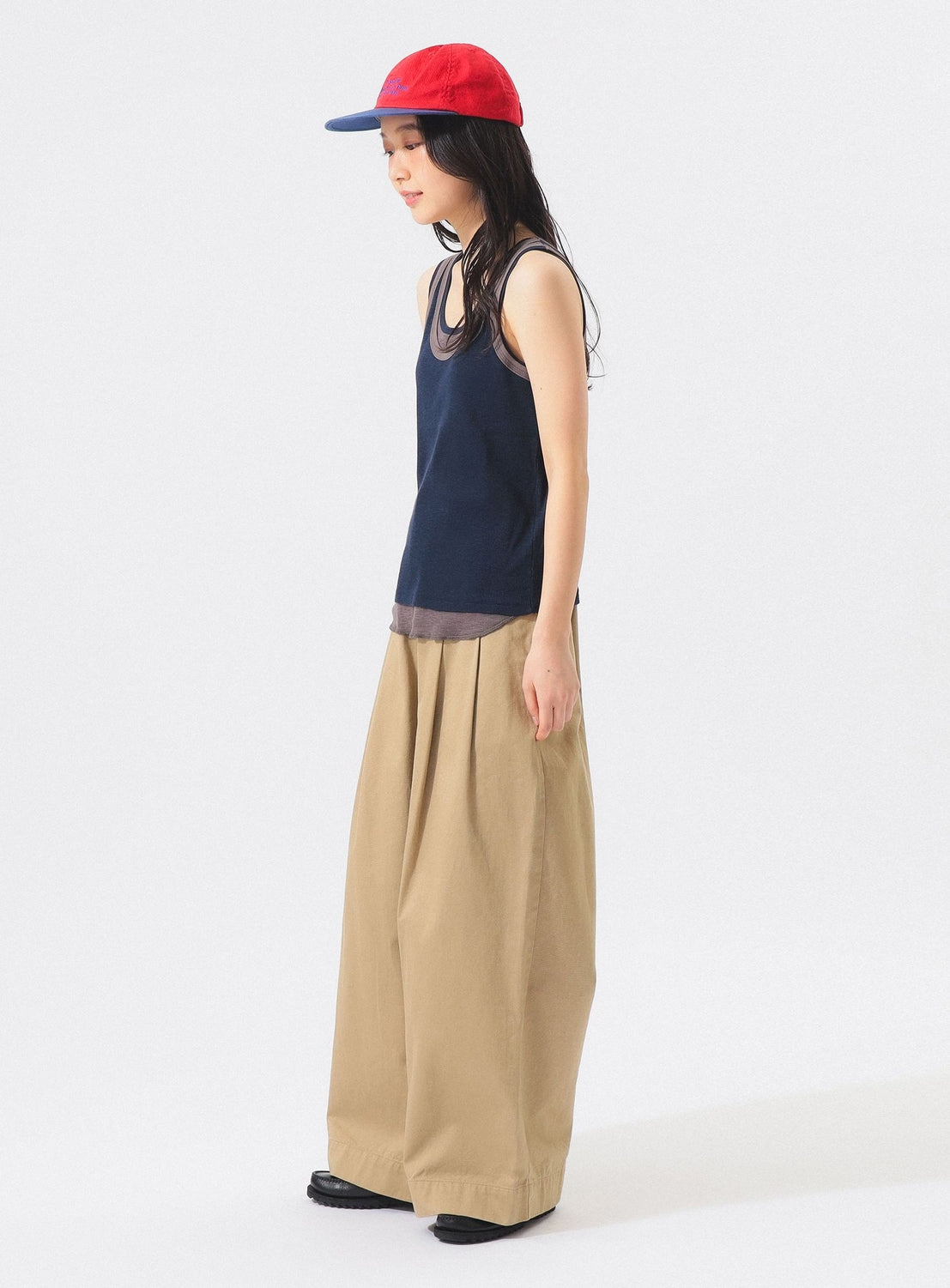 A person wearing the BEAMS BOY Layered Rib Tank Top in navy, along with a red cap and wide beige pants, stands against a plain white background, facing left.