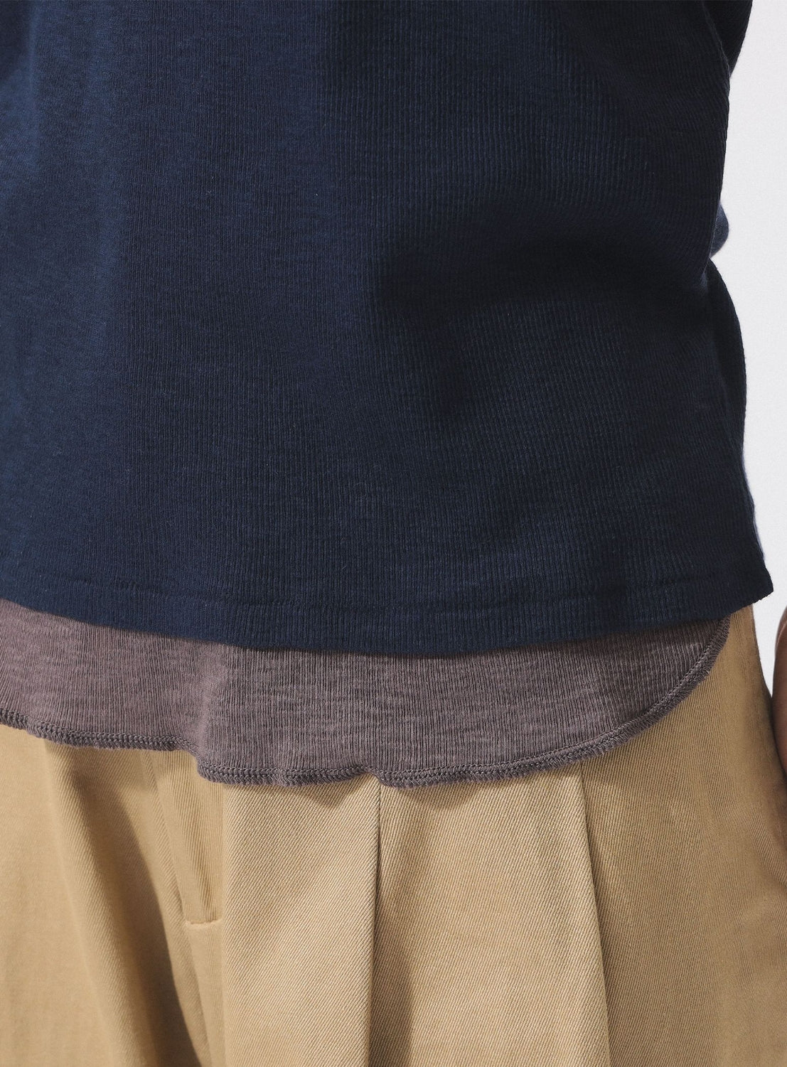 A person wearing a navy shirt layered over the BEAMS BOY Layered Rib Tank Top in brownish-grey, paired with tan pleated pants. Only the torso and upper legs are visible; the outfit highlights layered tops and neutral colors.