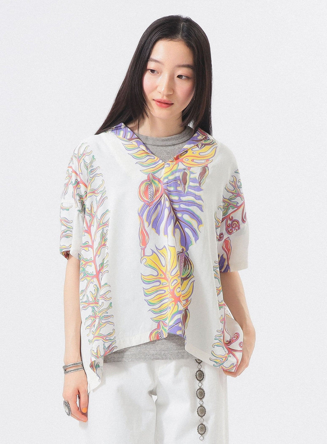 A woman with long dark hair wears the SUN SURF x BEAMS BOY Sailor Shirt, a loose white top with colorful Hawaiian prints, paired with light pants. She looks to the side and smiles slightly against a plain white background.