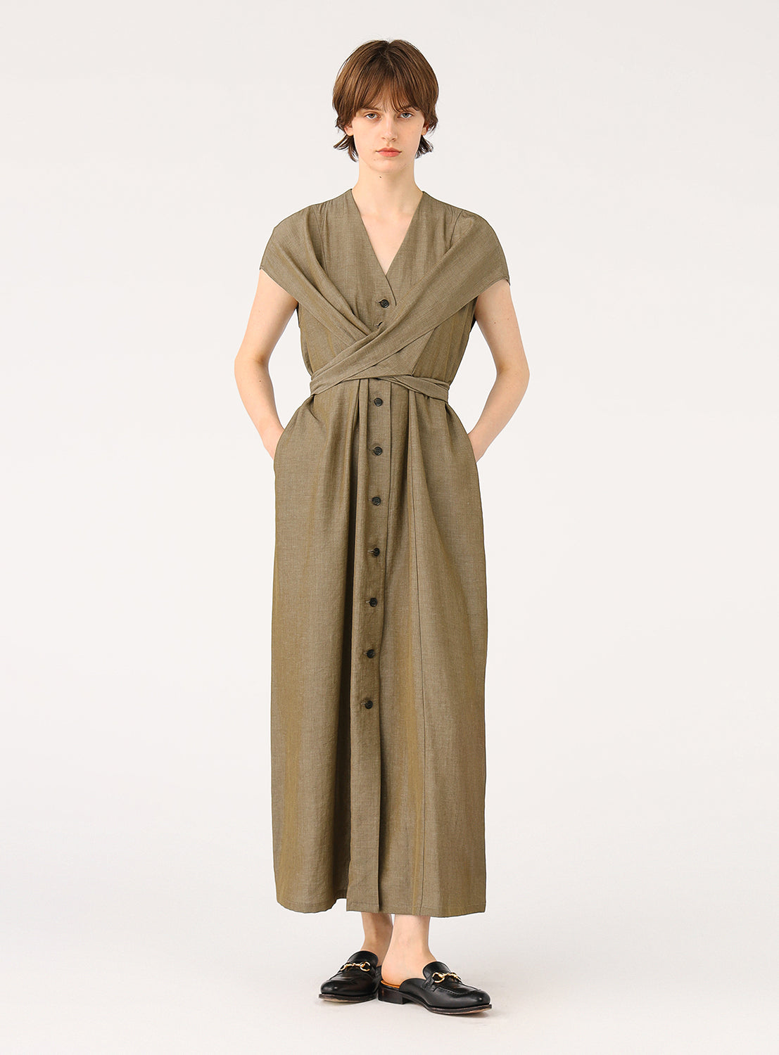 A female model wearing a long, sleeveless khaki dress with a V-neck and a tie waist, standing full body.