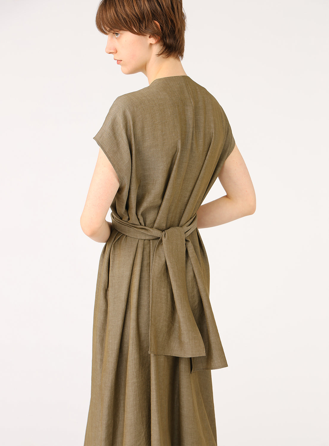  A female model from behind, wearing a long, sleeveless khaki dress, showing a tie detail at the back waist.