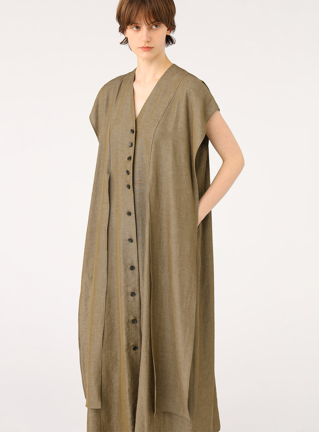 A female model wearing a long, sleeveless khaki dress, showing the V-neckline, front buttons, and a tie waist detail.