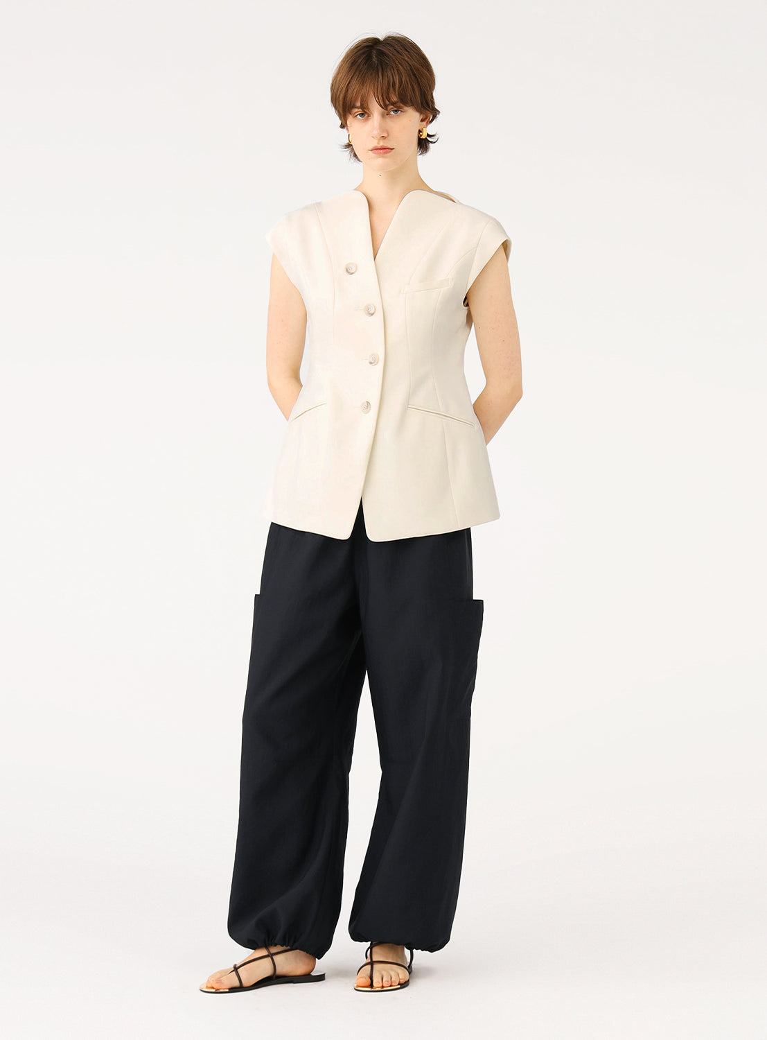 Female model in off-white tailored sleeveless vest and dark baggy pants, standing full body.