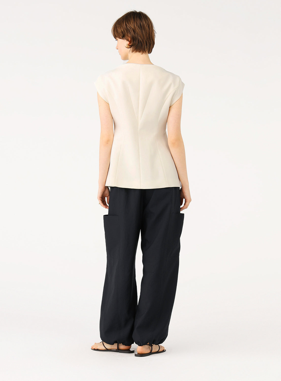 Female model from behind, wearing off-white tailored sleeveless vest and dark baggy pants.