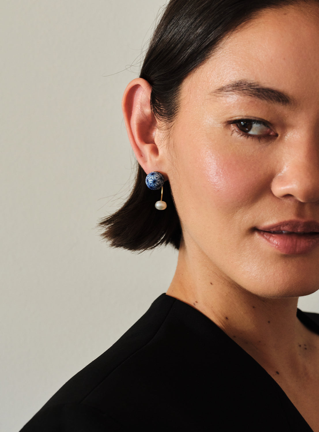 A woman with short dark hair wears a black top and striking Hizen Jewelry Arita Ware Dangling Motif & Pearl Earrings, reminiscent of Japanese handcrafted artistry. She is facing the camera with a soft, neutral expression against a plain light background.
