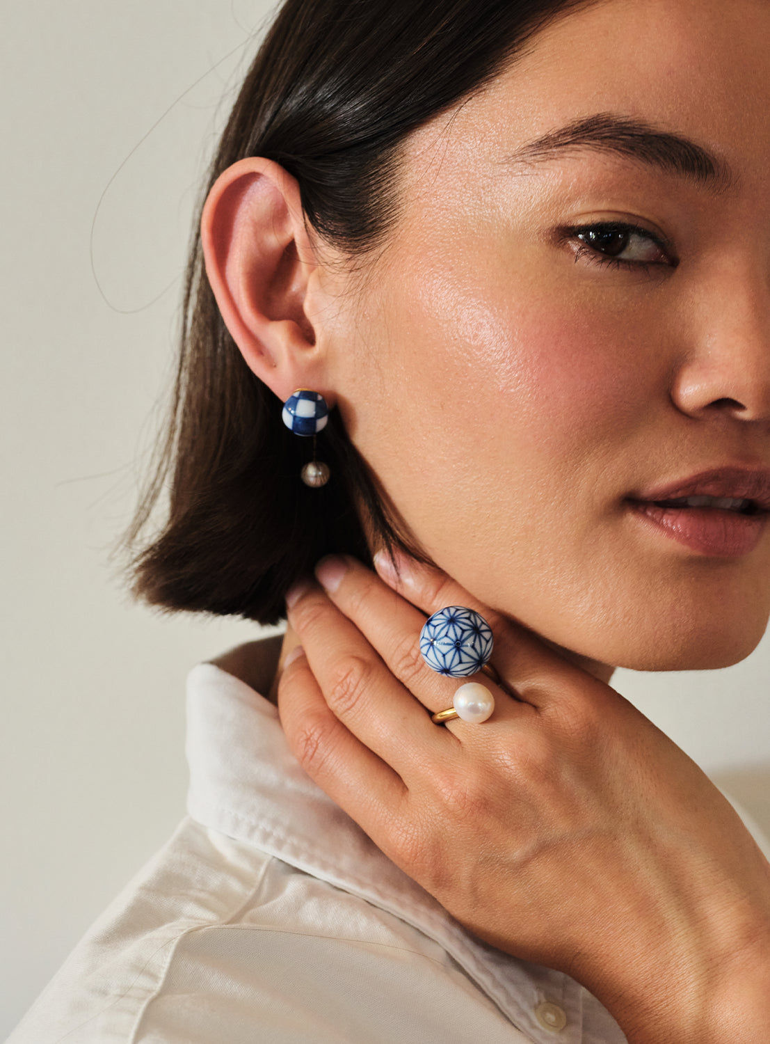 A woman with short dark hair wears a white shirt, accentuated by Hizen Jewelry’s Arita Ware Motif & Baroque Pearl Earrings—celebrating Japanese craftsmanship—and a matching ring adorned with freshwater pearls. Her hand rests gently near her jawline, drawing attention to the delicate jewelry.