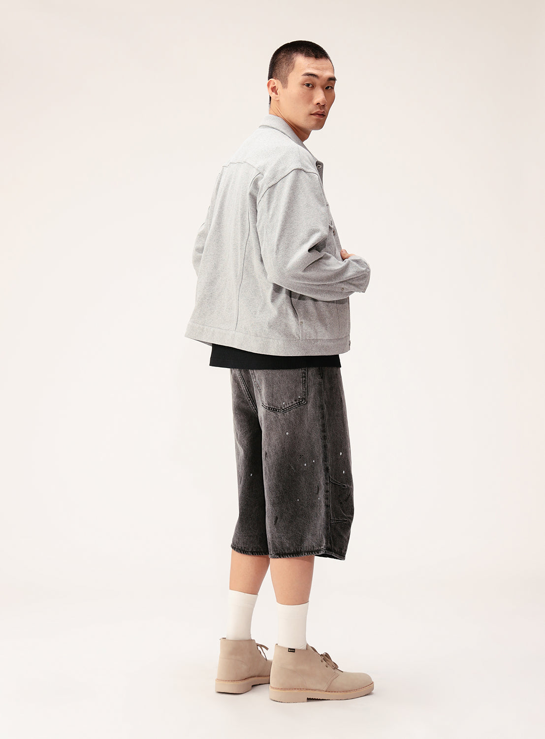 A young man stands sideways, glancing over his shoulder. He wears a BEAMS Button-Down Jersey Trucker Jacket with a black shirt, baggy gray knee-length shorts, white socks, and beige shoes against a plain light background.