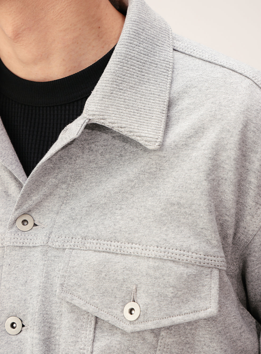 A close-up of a person wearing the BEAMS Button-Down Jersey Trucker Jacket in light gray over a black ribbed shirt, highlighting the collar, buttons, and part of the chest and neck.